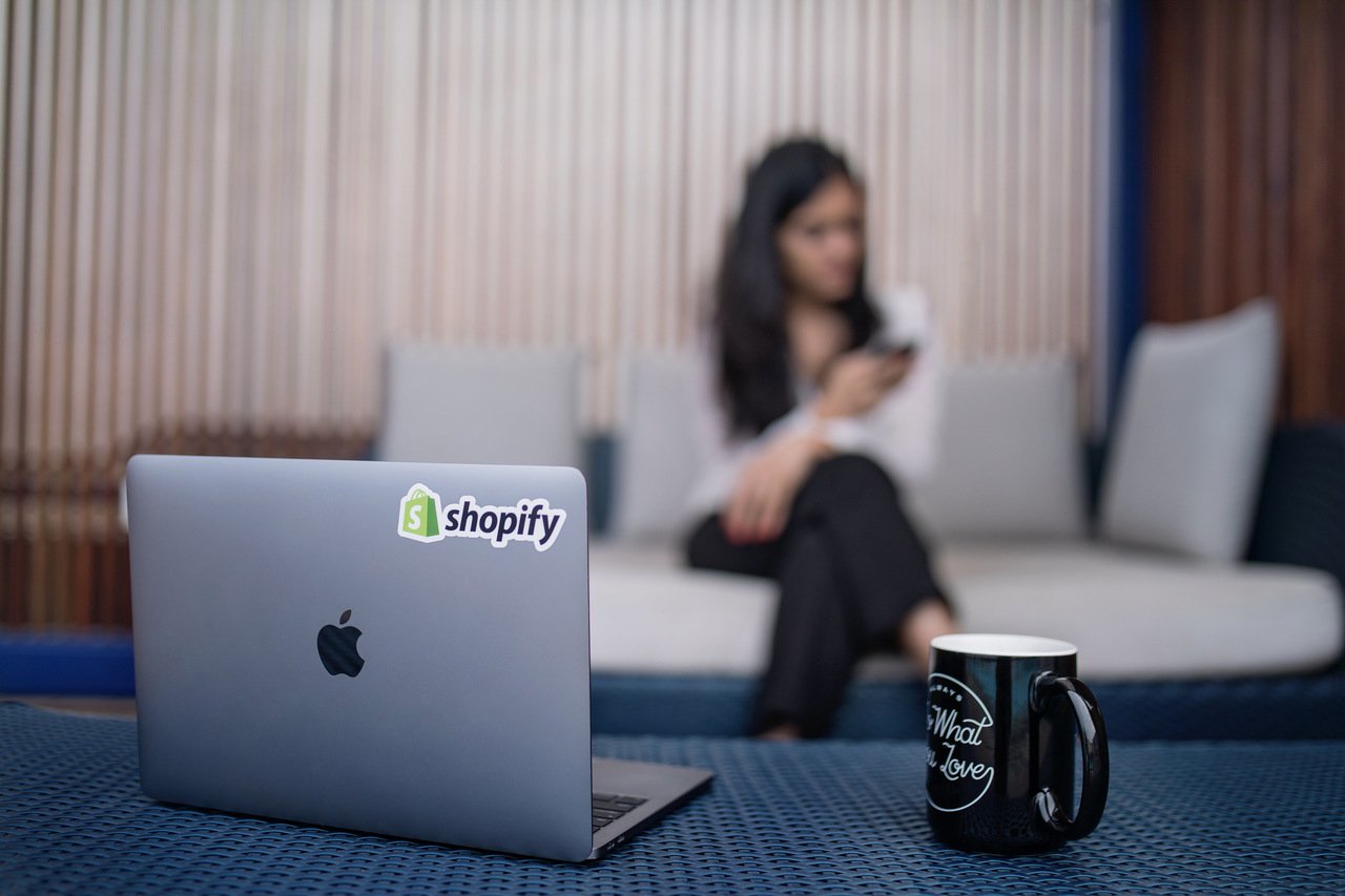 a MacBook with a Shopify sticker next to a mug and a woman sitting at the background