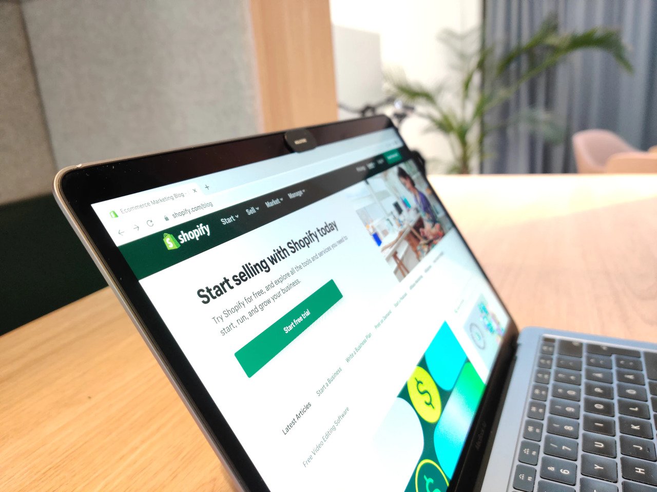 A stock photo showing a laptop with Shopify webpage on its screen, the laptop is on a table in a furnished room and a plant visible in the blurred background