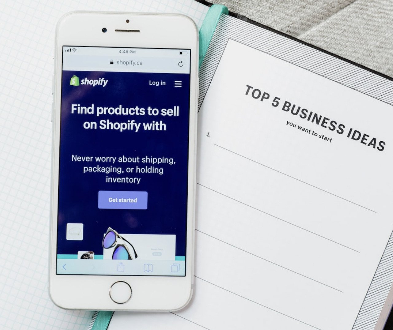 shopify stock photo showing a white phone with Shopify website open on the secreen on a notebook titled top 5 business ideas on it