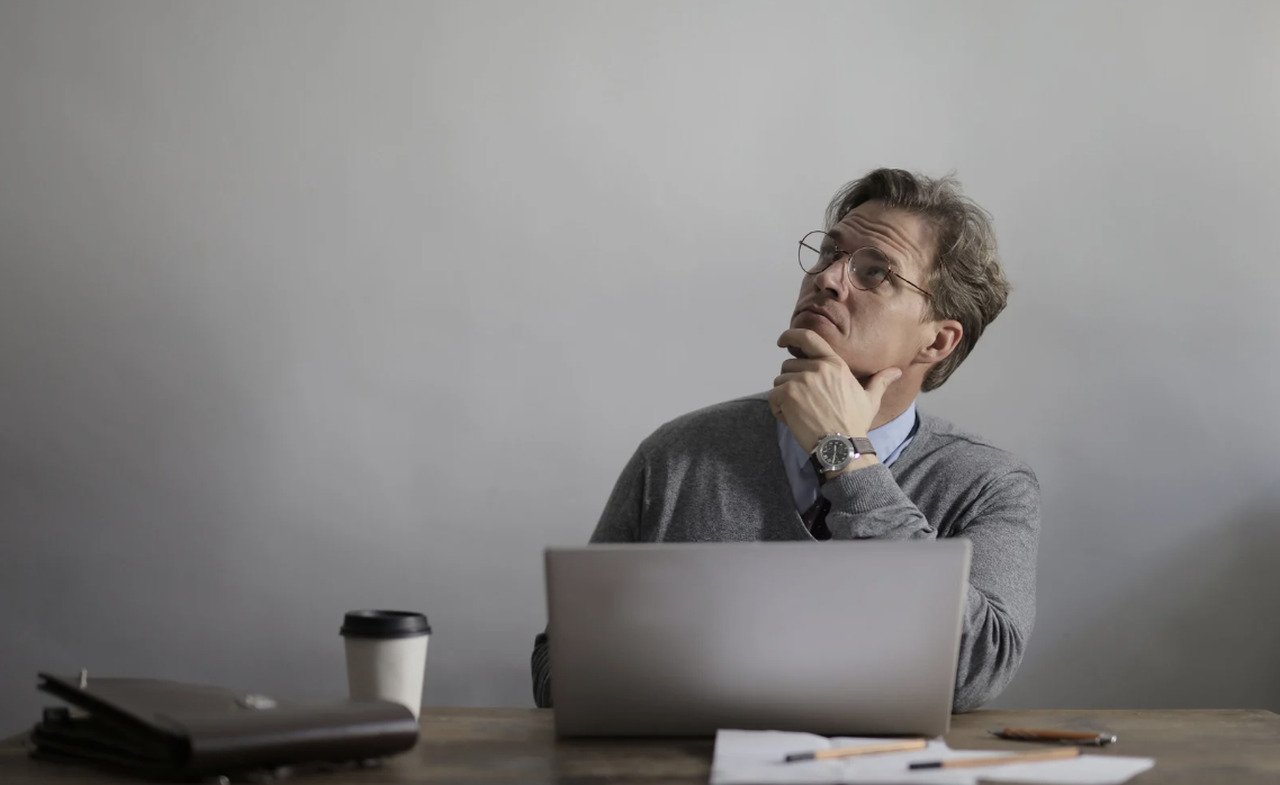 Man sitting behind a desk with a laptop and a cup of coffee holding his hand under his chin thinking and looking up