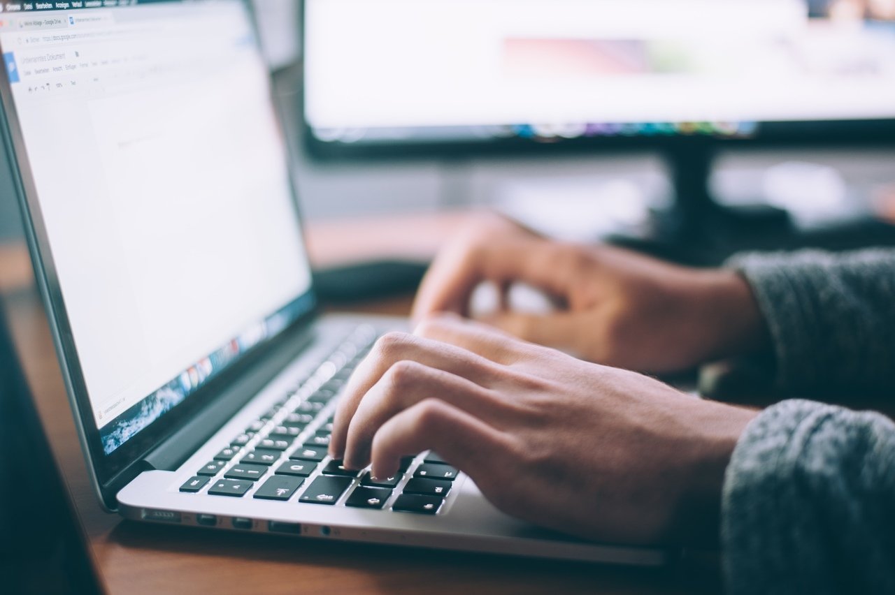 a person writing on a MacBook, only two hands and laptop are visible with a blurry background showing a computer screen