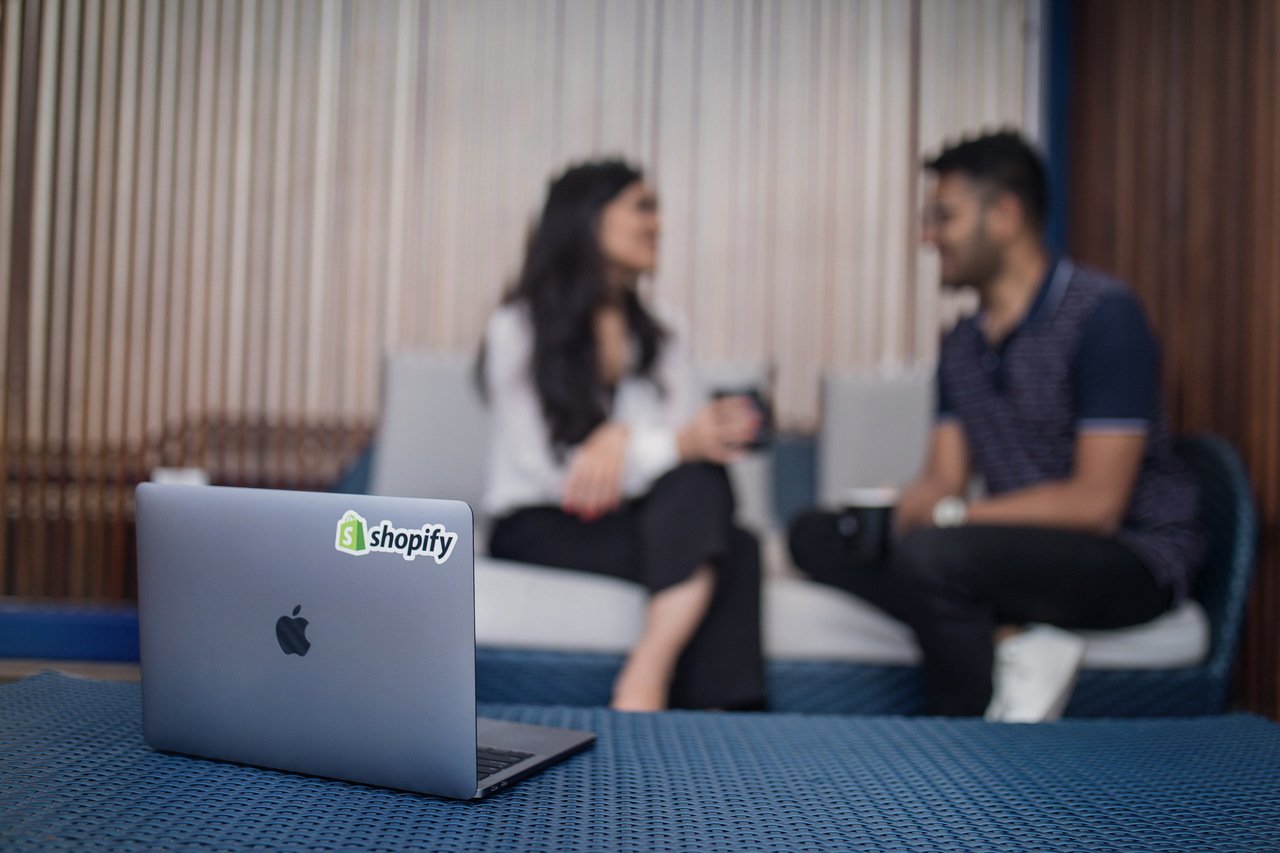 Shopify sticker on MacBook and two people talking at the background