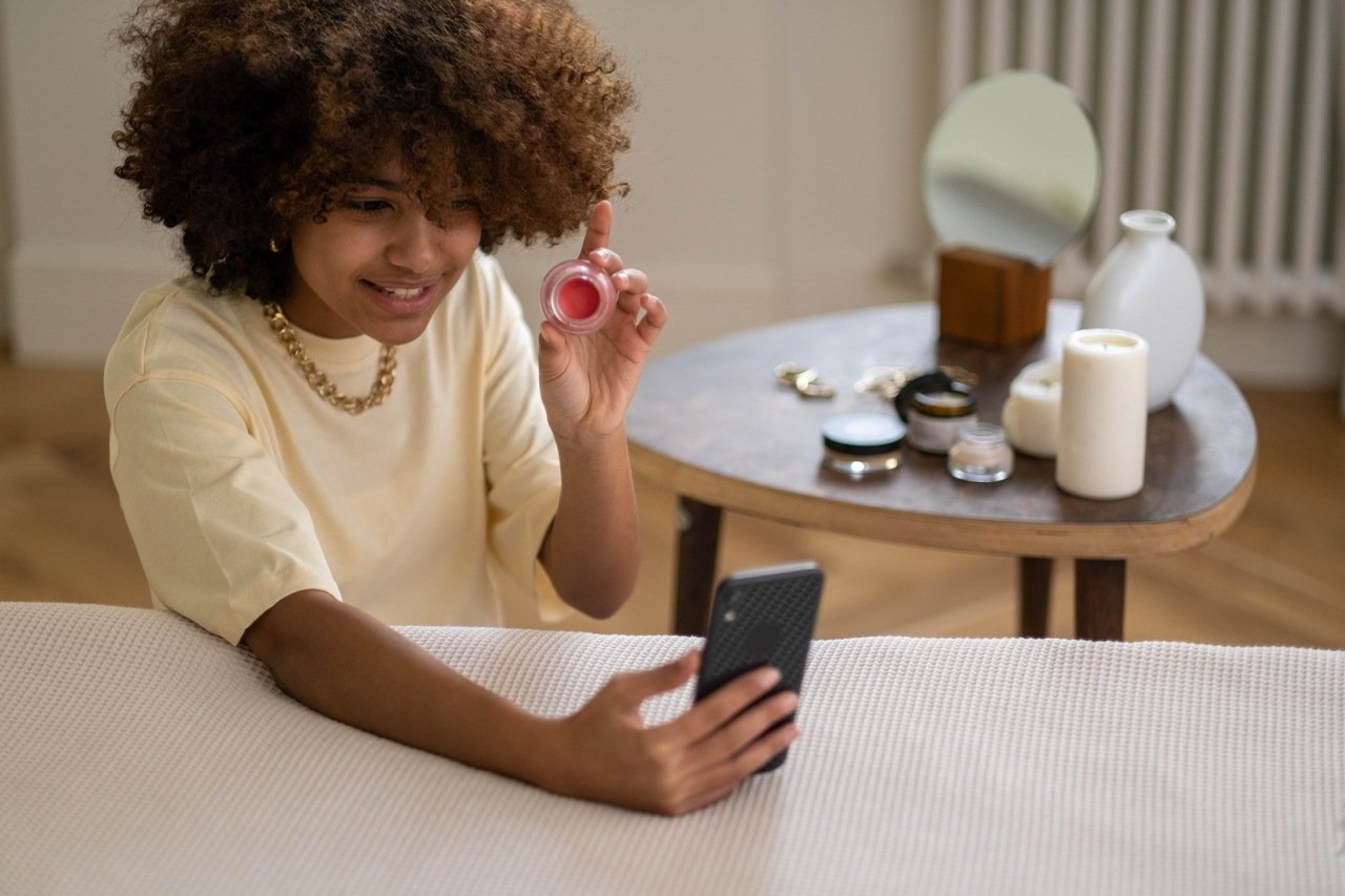 A curly haired woman holds a make-up product in her hand and demonstrates it on the phone.