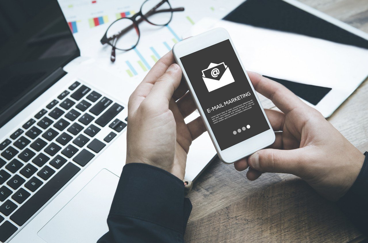 a hand holding an smart phone that shows the email marketing page in dark mode with a laptop and an eye glass on the background