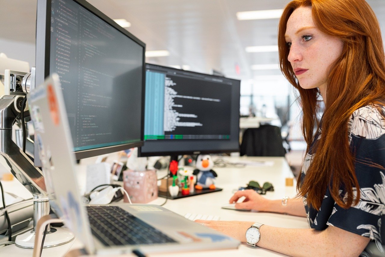 a ginger woman writing codes with two different monitors and a MacBook