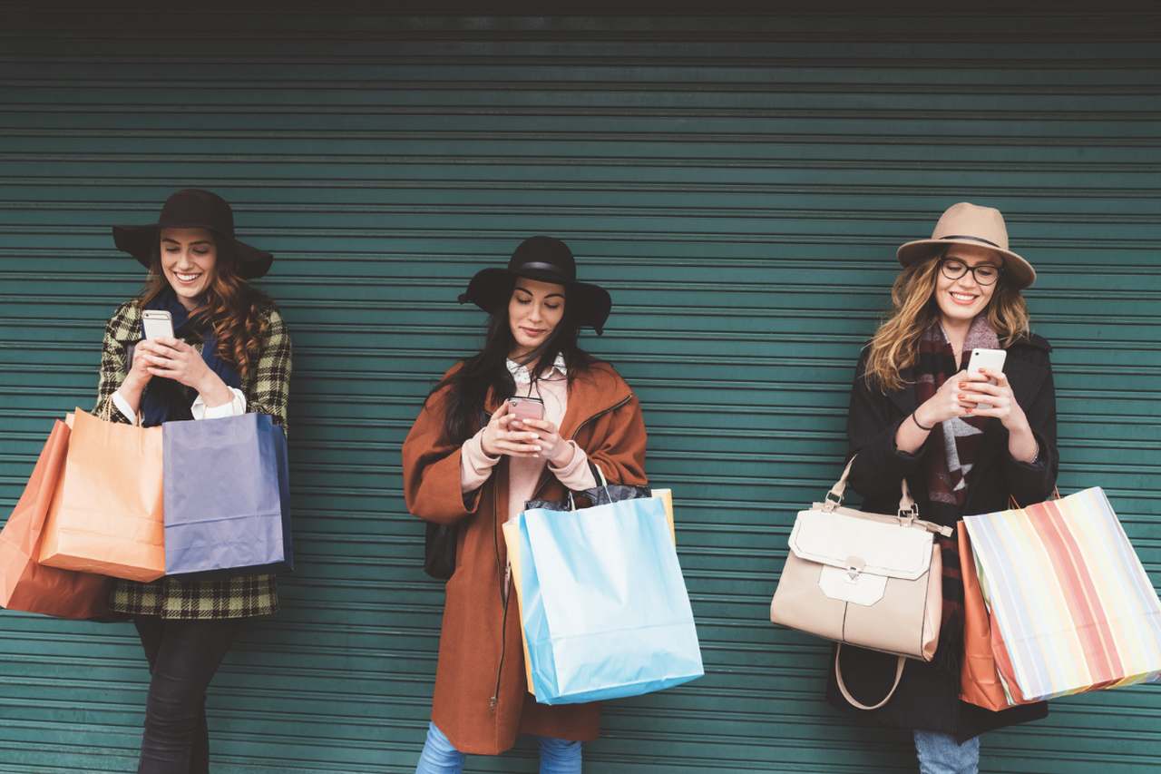 picture of three girls looking at their mobile phones smilling with shopping bags in their hands