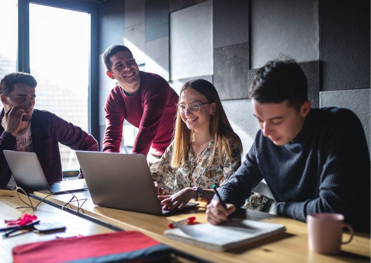 four people around the table checking computer and laughing with sincerity one is taking notes while one is giving advice