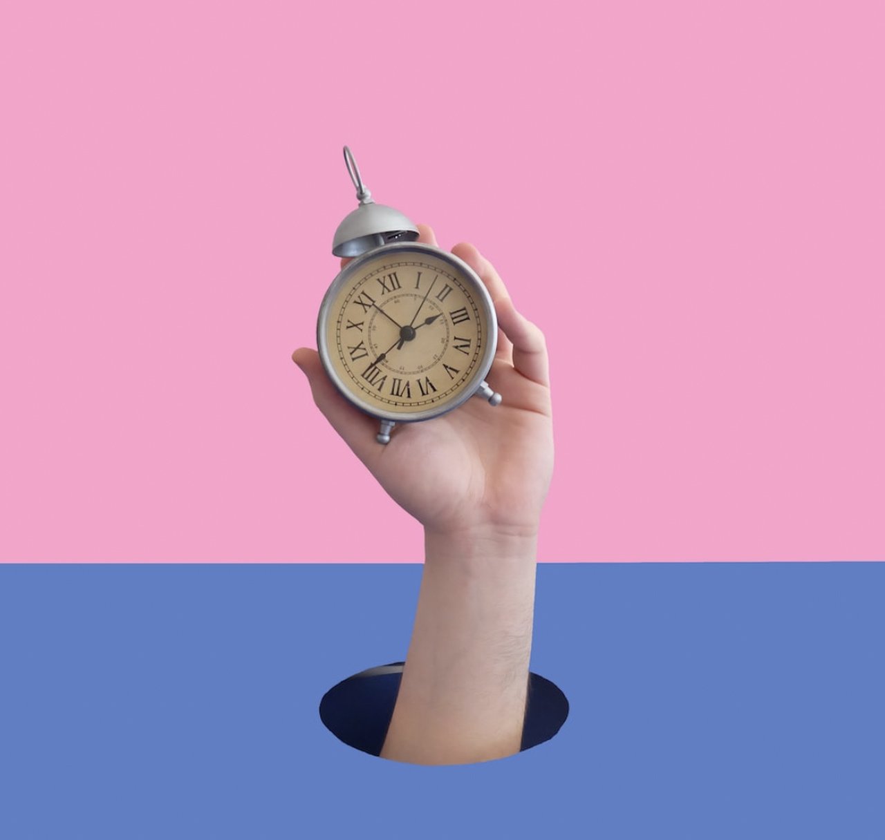 a hand holding a clock on a background split into two; the upper part is pink a nd the lower part is blue