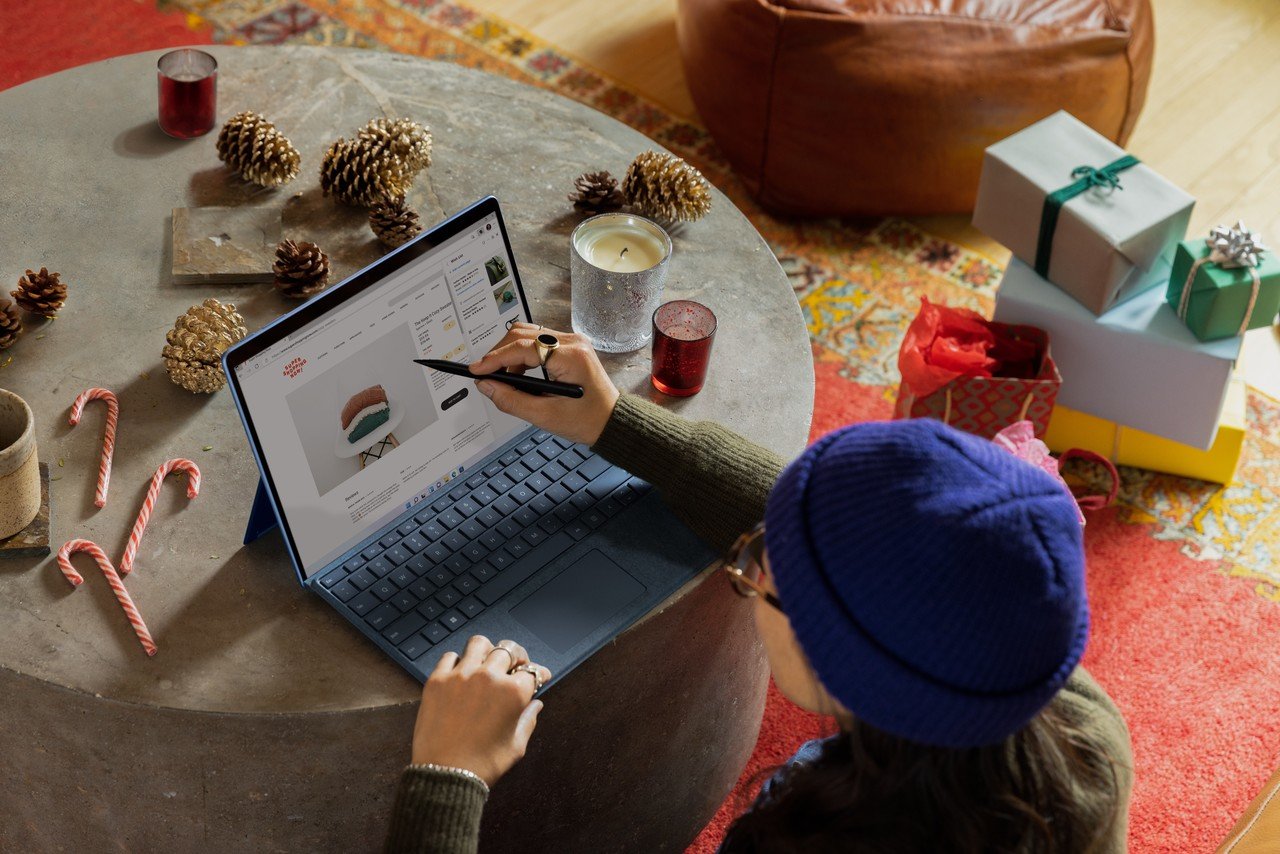 A person wearing an indigo hat, pointing at the laptop's screen at the round table; gift boxes are visible behind