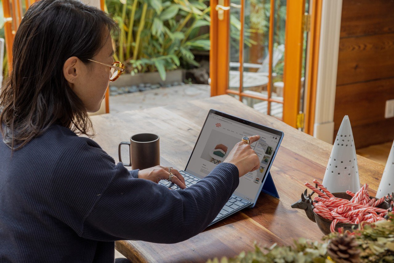 A person sitting at a desk in a room, pointing a finger on the screen of the laptop; a mug and some decoration stuff are on the table as well