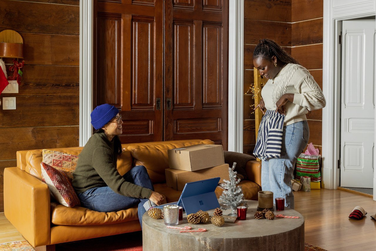 Two people, one male and one female, in a wooden wall spacious room, the woman is standing and showing the man her clothes; the man is sitting on a sofa with shopping bags