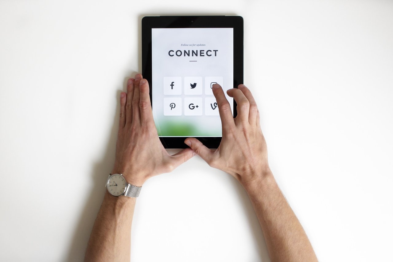 two hands holding a tablet at a white background; tablet has social media icons on the screen with a "connect" headline