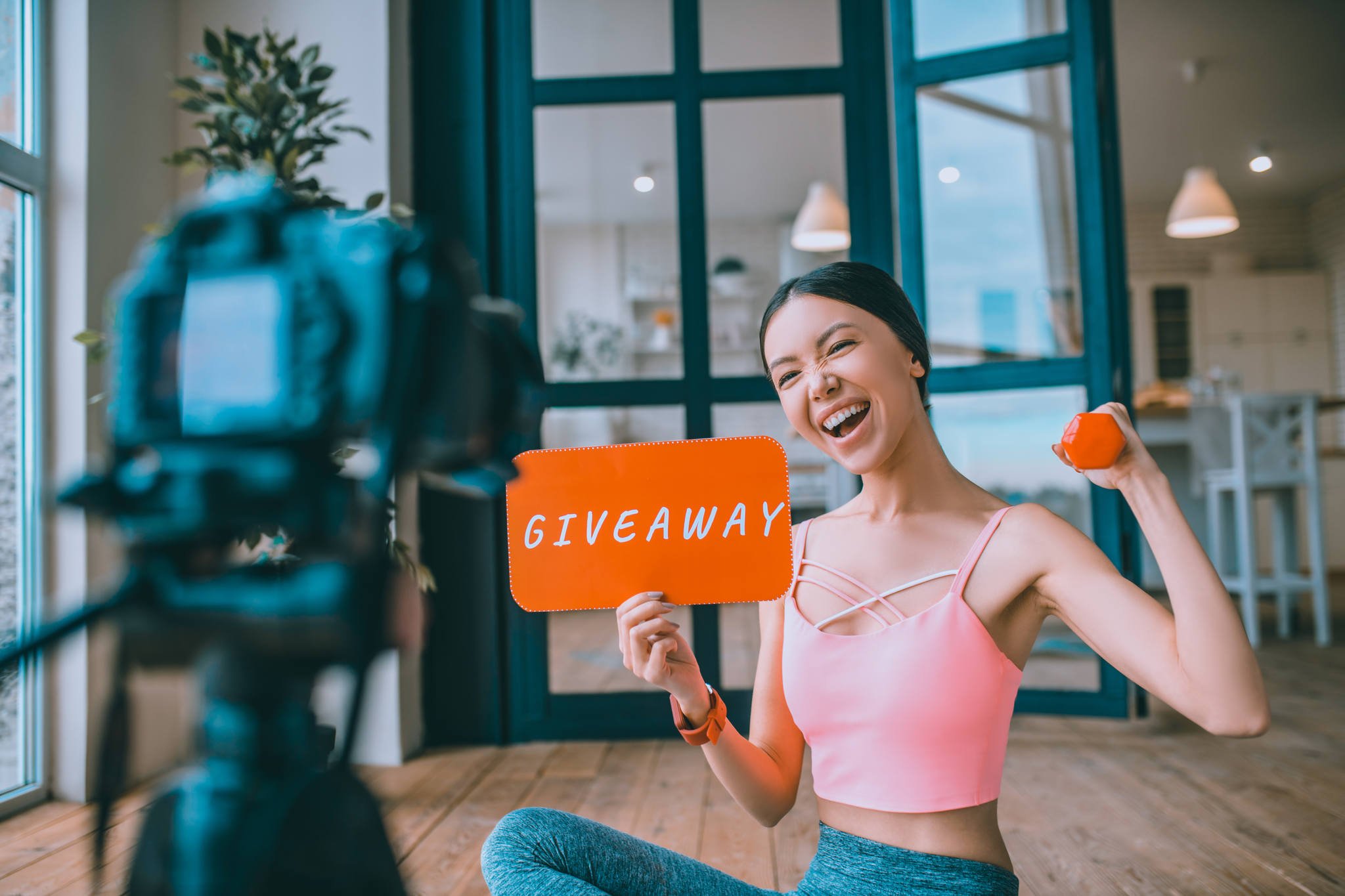 fitness blogger giveaway posing in front of a camera holding an orange cardboard written "giveaway while holding a dumbell on her other hand raising it on shoulder level