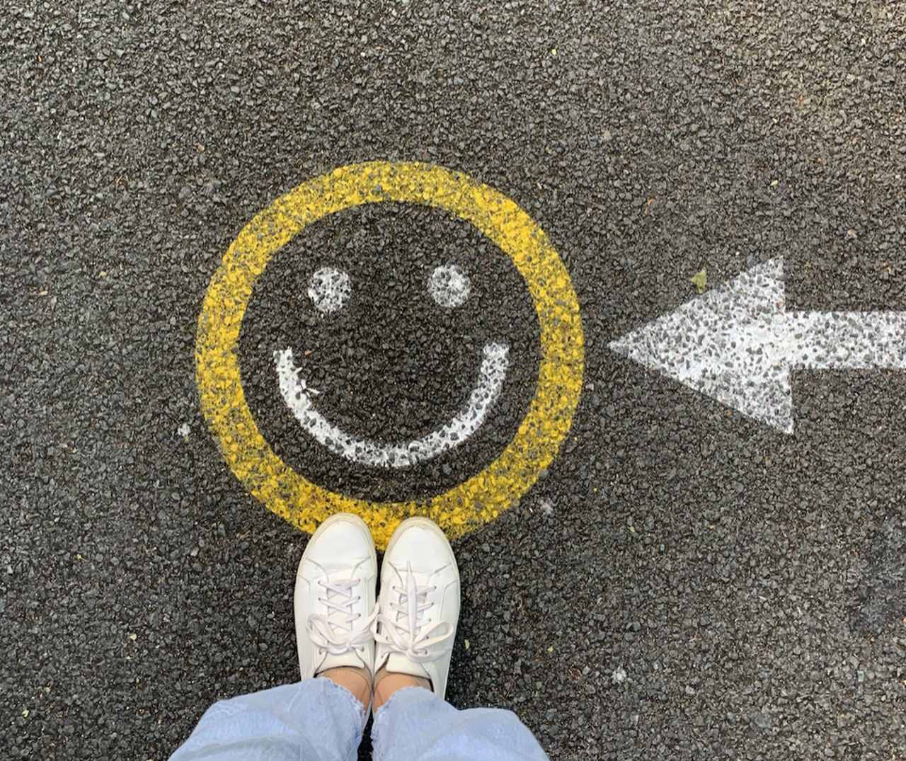 a circular happy face painted on the street with a white arrow pointing at it and white trainers of a person