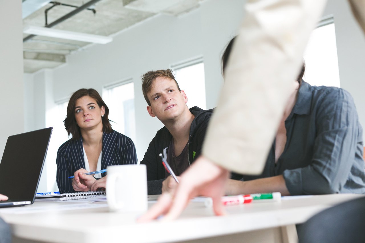 an arm is visible in the center tocuhing the table with fingertips, three other people are looking at the owner of the hand and one is taking notes in a meeting-room-like environment