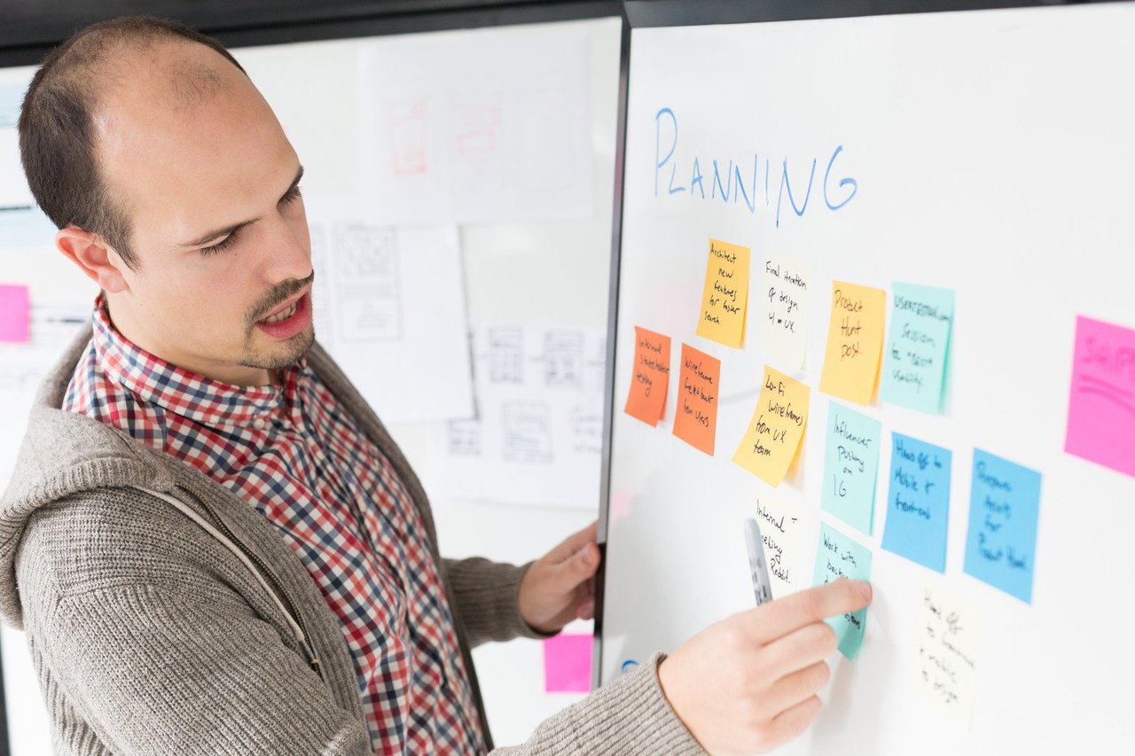 a man pointing on a whiteboard with sticky notes; his mouth is half open giving the impression that he is talking about the notes