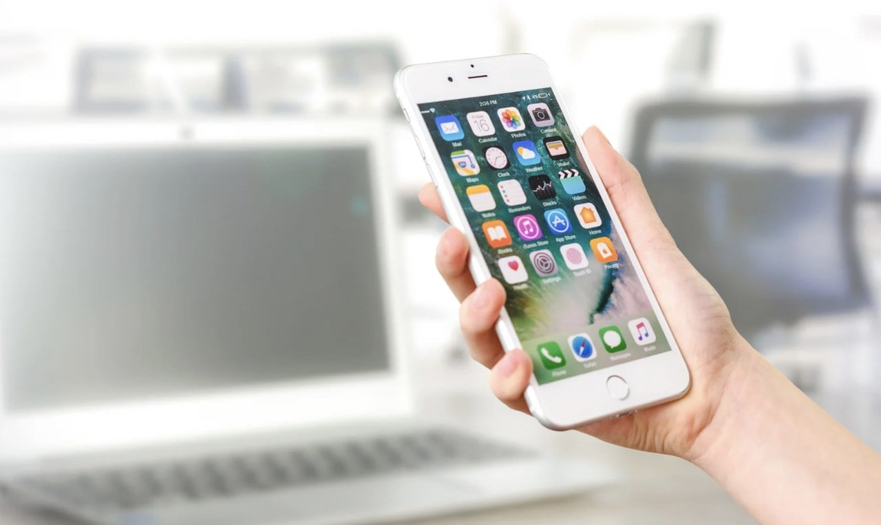 a hand holding a smartphone that shows all the apps installed on it; the background is blurry and a laptop is visible