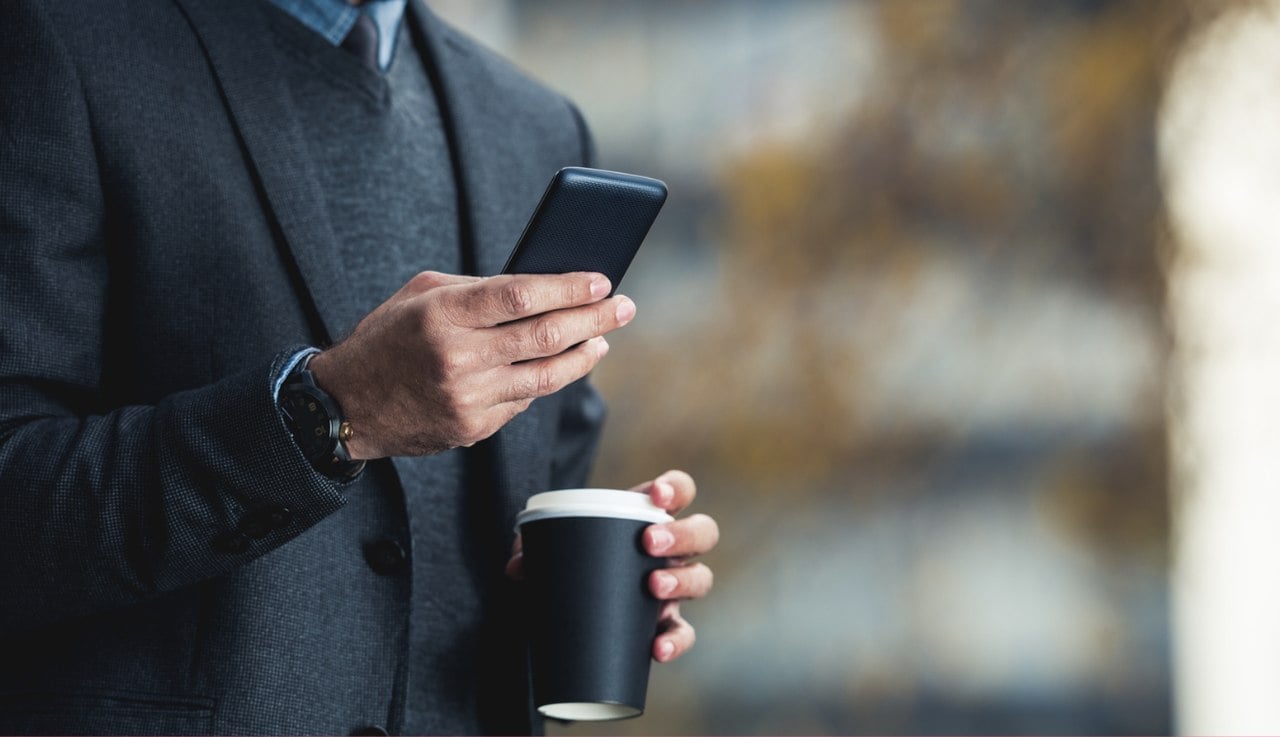 a business man in a black coat checking his phone with one hand and holding a cup of coffee with the other, standing on a blury background